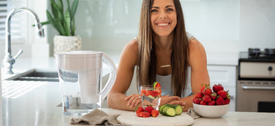 A woman smiling in the kitchen in front of a white Santevia MINA Alkaline Water Pitcher and some fresh fruits
