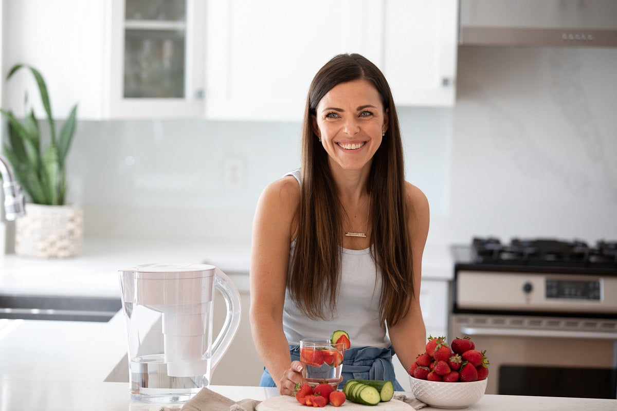 A smiling woman in a kitchen with a glass of strawberry and cucumber infused Santevia alkaline mineral water from her Santevia MINA Pitcher